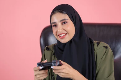Playing electronic games during the weekend, gestures of immersion and interaction while playing, using modern and advanced electronic technologies, a close-up portrait of an Arab Gulf Emirati woman wearing an abaya and hijab sitting on a chair holding a game controller, with a pink background.