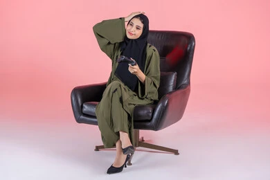 استخدام التقنيات الحديثة، قضاء اوقات ممتعة بلعب ألعاب الفيديو الترفيهية، تعابير الاحباط للخسارة، شابة سعودية تضع يدها على راسها بايماءات الحزن، بورتريه لإمرأة عربية خليجية اماراتية  تجلس على كرسي تحمل بيدها عصا التحكم، خلفية وردية