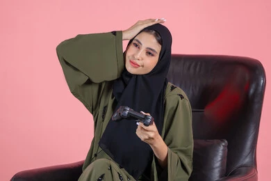 قضاء اوقات ممتعة بلعب ألعاب الفيديو الترفيهية، تعابير الاحباط للخسارة، استخدام التقنيات الحديثة، شابة سعودية تضع يدها على راسها بايماءات الحزن، بورتريه مقرب لإمرأة عربية خليجية اماراتية  تجلس على كرسي تحمل بيدها عصا التحكم، خلفية وردية