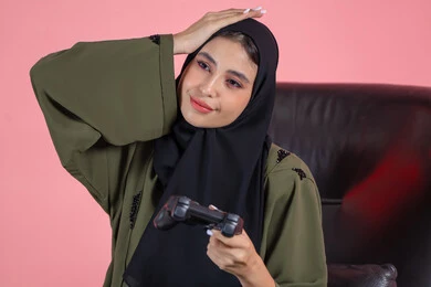 تعابير الاحباط للخسارة، استخدام التقنيات الحديثة، قضاء اوقات ممتعة بلعب ألعاب الفيديو الترفيهية، شابة سعودية تضع يدها على راسها بايماءات الحزن، بورتريه مقرب لإمرأة عربية خليجية اماراتية  تجلس على كرسي تحمل بيدها عصا التحكم، خلفية وردية