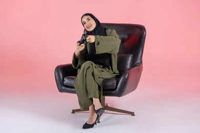 Enjoying video games during the weekend, gestures of immersion and interaction while playing, using modern and advanced electronic technologies, a portrait of an Arab Gulf Emirati woman wearing an abaya and hijab sitting on a chair holding a game controller, with a pink background.