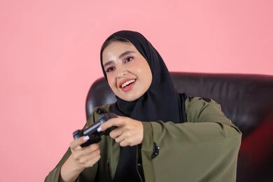 ممارسة العاب الفيديوز الترفيهية، ايماءات الاندماج والتفاعل أثناء اللعب، استخدام التقنيات الالكترونية الحديثة والمتطورة، بورتريه مقرب لإمرأة عربية خليجية إماراتية ترتدي العباءة و الحجاب تجلس على كرسي وتحمل بيدها عصا التحكم، خلفية وردية