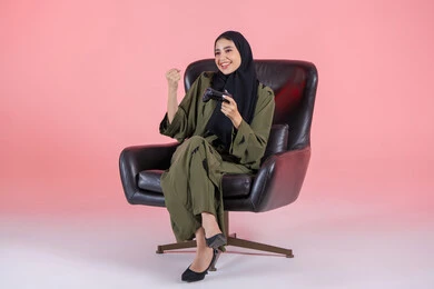 العالم الرقمي الترفيهي، الألعاب الالكترونية والأجهزة الحديثة ، ايماءات الفوز والانتصار، اللعب و التسلية بالعاب الفيديو ، بورتريه لإمرأة عربية خليجية اماراتية ترتدي العباءة و الحجاب تجلس على كرسي تمسك بيدها عصا التحكم , سيدة سعودية تنظر بتعابير السعادة ، الاستمتاع بتجربة ترفيهية مثيرة ،خلفية وردية