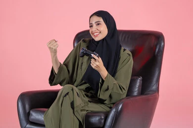 الألعاب الالكترونية والأجهزة الحديثة ، ايماءات الفوز والانتصار ، العالم الرقمي الترفيهي ، اللعب و التسلية بالعاب الفيديو ، بورتريه مقرب لإمرأة عربية خليجية اماراتية ترتدي العباءة و الحجاب تجلس على كرسي تمسك بيدها عصا التحكم , سيدة سعودية تنظر بتعابير السعادة ، الاستمتاع بتجربة ترفيهية مثيرة ،خلفية وردية