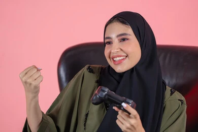 Gestures of victory and triumph, electronic games and modern devices, the entertaining digital world, playing and having fun with video games, a close-up portrait of an Emirati Gulf Arab woman wearing an abaya and hijab sitting on a chair holding a game controller, a Saudi woman looking with expressions of happiness, enjoying an exciting entertainment experience, pink background.
