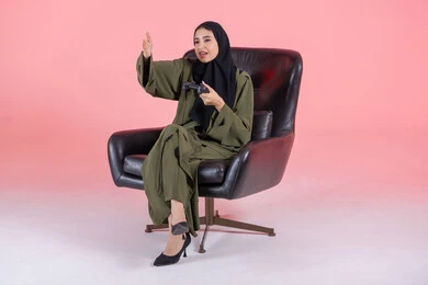 Spending free time in an enjoyable way, gestures and facial expressions indicating the difficulty of winning, entertaining modern techniques, a portrait of an Arab Gulf Emirati woman sitting on a chair holding a game controller and playing video games, with a pink background.