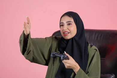 قضاء أوقات الفراغ باللعب بألعاب البلايستيشن الترفيهية، إيماءات وتعابير وجه ويدين تدل على صعوبة الفوز، تقنيات حديثة مسلية ، بورتريه لإمرأة عربية خليجية اماراتية تجلس على كرسي تحمل بيدها عصا التحكم و تلعب بألعاب الفيديو، خلفية وردية
