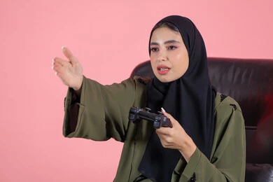 تقنيات حديثة مسلية, إيماءات وتعابير وجه ويدين تدل على صعوبة الفوز, قضاء أوقات الفراغ باللعب بألعاب البلايستيشن الترفيهية ، بورتريه لإمرأة عربية خليجية اماراتية تجلس على كرسي تحمل بيدها عصا التحكم و تلعب بألعاب الفيديو، خلفية وردية