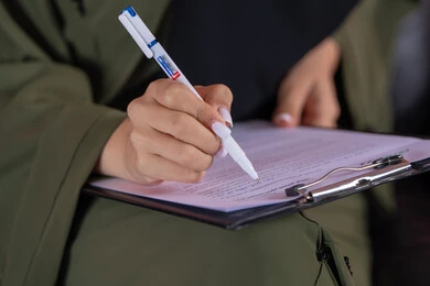 استخدام الورق الأبيض في تسجيل الملاحظات والمعلومات المهمة، مفهوم كتابه وتدوين الملاحظات، بورتريه مقرب ليد امراة عربية خليجية اماراتية تمسك بيدها قلم تقوم بالكتابة على نوت، خلفية وردية