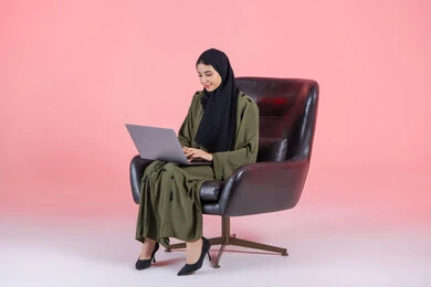 Integrating technology into work, accomplishing required tasks via laptop, managing businesses remotely, using modern and advanced devices, a portrait of an Emirati Gulf Arab woman wearing an abaya and hijab sitting on a chair using a laptop, women's professions and jobs, pink background.
