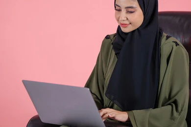 Remote business management, using modern and advanced devices, a close-up portrait of an Emirati Gulf Arab woman wearing an abaya and hijab sitting on a chair using a laptop, integrating technology into work, completing tasks required via the laptop, women's professions and jobs, pink background.