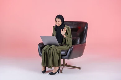 Spending enjoyable time, watching a movie on a mobile device, making video calls online, playing on the laptop, managing projects remotely, raising the hand with gestures of peace and greeting, a portrait of an Arab Gulf Emirati woman wearing an abaya and hijab sitting on a chair completing work tasks on the computer, entertainment and amusement, pink background.