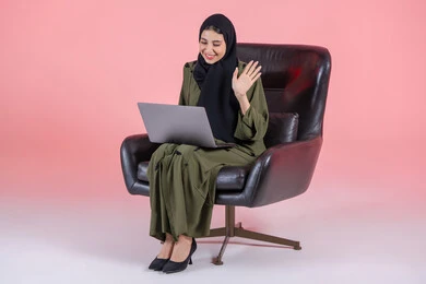 مشاهدة فلم عبر الجهاز المحمول، إجراء مكالمات فيديو عبر الإنترنت، اللعب على اللابتوب، إدارة المشاريع عن بعد، رفع كف الايد بايماءات السلام و التحيه، بورتريه لامرأة عربية خليجيه اماراتية ترتدي العباءة و الحجاب تجلس على كرسي تقوم بانجاز مهام العمل عبر الحاسوب, تسليه وترفيه, قضاء وقت ممتع، خلفية وردية