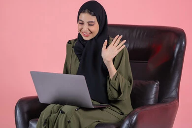 Making video calls online, playing on the laptop, managing projects remotely, raising the hand with gestures of peace and greeting, a portrait of an Emirati Gulf Arab woman wearing an abaya and hijab sitting on a chair completing work tasks on the computer, watching a movie on a mobile device, entertainment and leisure, having a good time, pink background.