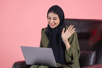 اللعب على اللابتوب، إدارة المشاريع عن بعد، رفع كف الايد بايماءات السلام و التحيه، بورتريه لامرأة عربية خليجيه اماراتية ترتدي العباءة و الحجاب تجلس على كرسي تقوم بانجاز مهام العمل عبر الحاسوب, إجراء مكالمات فيديو عبر الإنترنت, مشاهدة فلم عبر الجهاز المحمول, تسليه وترفيه, قضاء وقت ممتع، خلفية وردية