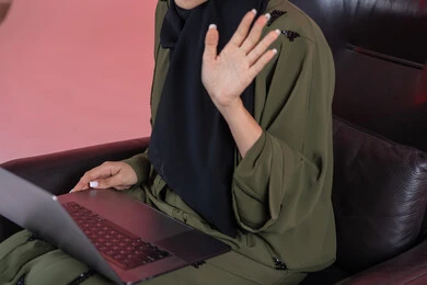 Remote project management, raising the hand in gestures of peace and greeting, a close-up portrait of an Emirati Gulf Arab woman wearing an abaya sitting on a chair completing work tasks on the computer, playing on the laptop, making video calls online, watching a movie on a mobile device, entertainment and leisure, having a good time, pink background.