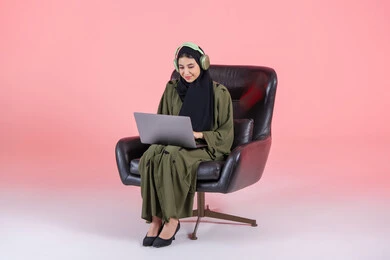 Executing the required tasks via a laptop, women's professions and jobs, integrating technology into work, using modern and advanced devices, managing business remotely, a portrait of an Emirati Gulf Arab woman wearing a hijab and abaya with headphones sitting on a chair using a laptop, pink background.