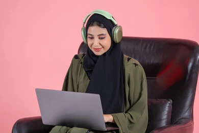 إنجاز المهام المطلوبة عبر اللابتوب، دمج التكنولوجيا بالعمل، استخدام الأجهزة الحديثة والمتطورة، ادارة الأعمال عن بعد، بورتريه لامرأة عربية خليجيه اماراتية ترتدي الحجاب و العباءة و سماعات الرأس  تجلس على كرسي تقوم باستخدام الحاسوب المحمول، مهن ووظائف نسائية، خلفية وردية