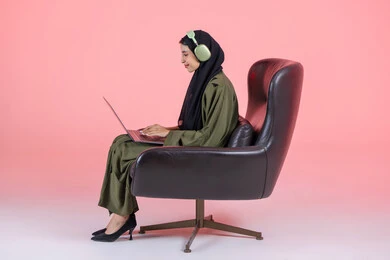 دمج التكنولوجيا بالعمل، استخدام الأجهزة الحديثة والمتطورة، ادارة الأعمال عن بعد، بورتريه من الجانب لامرأة عربية خليجيه اماراتية ترتدي الحجاب و العباءة و سماعات الرأس  تجلس على كرسي تقوم باستخدام الحاسوب المحمول، إنجاز المهام المطلوبة عبر اللابتوب، مهن ووظائف نسائية، خلفية وردية