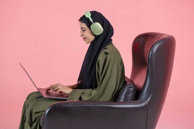 The use of modern and advanced devices, remote business management, a side portrait of an Emirati Gulf Arab woman wearing a hijab and abaya with headphones sitting on a chair using a laptop, integrating technology with work, completing required tasks via the laptop, women's professions and jobs, pink background.