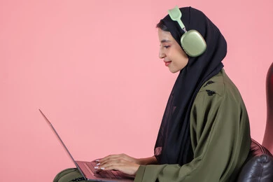 Remote business management, using modern and advanced devices, a side portrait of an Emirati Gulf Arab woman wearing a hijab and abaya with headphones sitting on a chair using a laptop, integrating technology with work, completing required tasks via the laptop, women's professions and jobs, pink background.