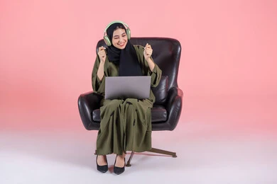 Modern technical devices, interaction with musical tones, a Saudi woman enjoying her time listening to songs, the concept of listening and using headphone technologies, a portrait of an Arab Gulf Emirati woman wearing a hijab and abaya with headphones sitting on a chair with a laptop on her lap, pink background.