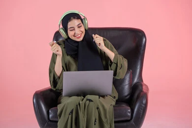 التفاعل مع النغمات الموسيقية، سيدة سعودية تقوم بقضاء أوقات ممتعة بسماع الأغاني، مفهوم الإصغاء واستخدام تقنيات الهيدفون، بورتريه لامرأة عربية خليجية اماراتية ترتدي الحجاب والعباءة وسماعات الرأس تجلس على كرسي تضع الحاسب المحمول على ساقيها، الأجهزة التقنية الحديثة، خلفية وردية