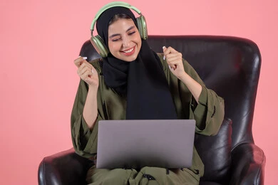 Psychological integration with musical rhythms, a Saudi woman enjoying her time listening to songs, the concept of listening and using headphone techniques, a portrait of an Arab Gulf Emirati woman wearing a hijab and abaya with headphones sitting on a chair with a laptop on her lap, modern technological devices, pink background.