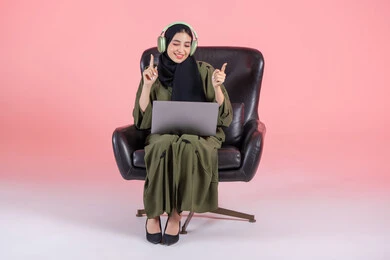 Enjoying musical tunes and interacting with them, a Saudi woman spends enjoyable times listening to songs, the concept of listening and using headphone technologies, a portrait of an Arab Gulf Emirati woman wearing a hijab and abaya with headphones sitting on a chair with a laptop on her lap, modern technological devices, pink background.