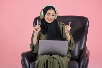 التفاعل مع النغمات الموسيقية والشعور بالإيقاع، سيدة سعودية تقوم بقضاء أوقات ممتعة بسماع الأغاني، مفهوم الإصغاء واستخدام تقنيات الهيدفون، بورتريه لامرأة عربية خليجية اماراتية ترتدي الحجاب والعباءة وسماعات الرأس تجلس على كرسي تضع الحاسب المحمول على ساقيها، الأجهزة التقنية الحديثة، خلفية وردية