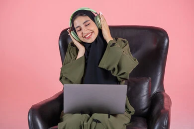 الاستمتاع بالموسيقى والانغماس في الإيقاعات، سيدة سعودية تقوم بقضاء أوقات ممتعة بسماع الأغاني، مفهوم الإصغاء واستخدام تقنيات الهيدفون، بورتريه لامرأة عربية خليجية اماراتية ترتدي الحجاب والعباءة وسماعات الرأس تجلس على كرسي تضع الحاسب المحمول على ساقيها، الأجهزة التقنية الحديثة، خلفية وردية