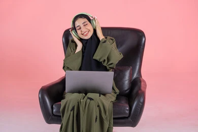 A Saudi woman is spending enjoyable time listening to music, immersing herself in the musical tunes, the concept of listening, and using headphone technology. A portrait of an Arab Gulf Emirati woman wearing a hijab and abaya with headphones sitting on a chair, placing a laptop on her lap, modern technological devices, with a pink background.