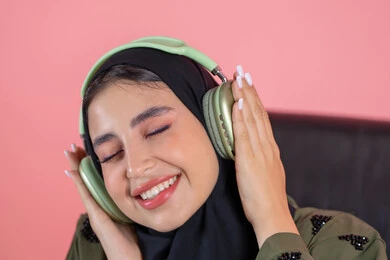 The concept of listening and using headphone techniques, merging with musical tunes, a Saudi woman enjoying her time listening to songs, a close-up portrait of an Emirati Gulf Arab woman wearing a hijab and abaya with headphones, modern technological devices, pink background.