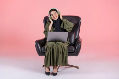 الاندماج مع الأنغام الموسيقية، مفهوم الإصغاء واستخدام تقنيات الهيدفون، سيدة سعودية تقوم بقضاء أوقات ممتعة بسماع الأغاني، بورتريه لامرأة عربية خليجية اماراتية ترتدي الحجاب والعباءة وسماعات الرأس تضع الحاسب المحمول على ساقيها، الأجهزة التقنية الحديثة، خلفية وردية