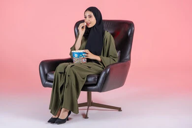 Snacking and snacks, home entertainment activities, having a good time at home, gestures of engagement and focus, a portrait of an Emirati Gulf Arab girl wearing a hijab and abaya sitting on a chair holding a box of crunchy popcorn, enjoying watching movies and series, following TV programs, pink background.
