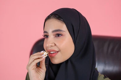 ايماءات الاندماج و التركيز، قضاء وقت ممتع في المنزل، انشطة ترفيهية منزلية، تناول الوجبات الخفيفة والسناك، بورتريه مقرب لفتاة عربية خليجية اماراتية ترتدي الحجاب تحمل بيديها علبة من الفوشار المقرمش، الاستمتاع بمشاهدة الأفلام والمسلسلات، متابعة برامج التلفاز، خلفية وردية