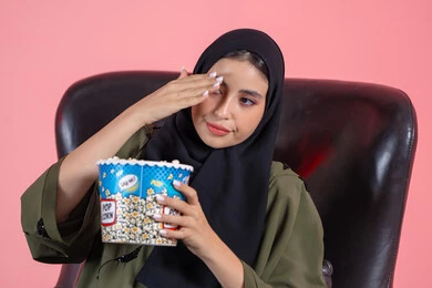 Spending enjoyable and entertaining times, watching an emotional drama film, feeling emotions while following drama films, a close-up portrait of an Arab Gulf Emirati woman wearing a hijab and an abaya sitting on a chair holding a bucket of popcorn in her hands, with a pink background. Spending enjoyable and entertaining times, watching an emotional drama film, feeling emotions while following drama films, a close-up portrait of an Arab Gulf Emirati woman wearing a hijab and an abaya sitting on a chair holding a bucket of popcorn in her hands, with a pink background.