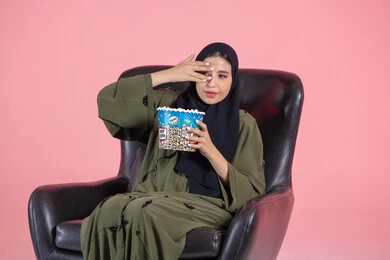 مفهوم التسلية والترفيه، تعابير الاندماج و التأثر، مشاهدة الأفلام والمشاهد الدرامية المؤثرة، بورترية  لامراة عربية خليجية اماراتية محجبة ترتدي العباءة تجلس على الكرسي تضع يديها على عينها  تحمل بيدها كوب مليء بالفوشار، خلفية وردية