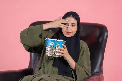 تناول الفشار اثناء مشاهدة الموفي، بورترية مقرب لفتاه عربيه خليجيه اماراتية ترتدي الحجاب تجلس علي كرسي تضع يدها على وجهها وتنظر من بين اصابعها اثناء مشاهدة فلم مخيف، ايماءات الخوف والتوتر، خلفية وردية