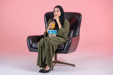 ايماءات الاندماج و التركيز، متابعة المسلسلات والبرامج التلفزيونية، تناول العصائر الباردة والسناكات الخفيفة، بورتريه لفتاة عربية خليجية اماراتية ترتدي الحجاب و العباءة تجلس على كرسي تحمل كأس العصير والفشار،قضاء أوقات ممتعة، خلفية بيضاء