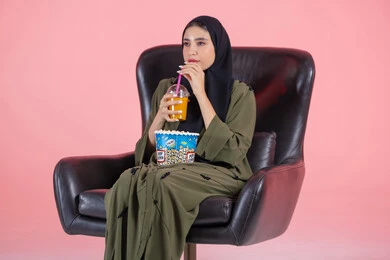 متابعة المسلسلات والبرامج التلفزيونية، ايماءات الاندماج و التركيز، تناول العصائر الباردة والسناكات الخفيفة، بورتريه لفتاة عربية خليجية اماراتية ترتدي الحجاب و العباءة تجلس على كرسي تحمل كأس العصير والفشار،قضاء أوقات ممتعة، خلفية بيضاء
