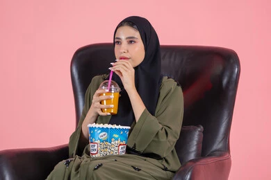 الاستمتاع بمشاهدة الأفلام والمسلسلات، تناول الفوشار والمشروبات الباردة، قضاء أوقات ممتعة، بورتريه لامرأة اماراتية عربية خليجية محجبة ترتدي عباءة خضراء تجلس على كرسي مريح و تشرب مشروب منعش و تضع دلو الفشار على ساقيها, خلفية وردية