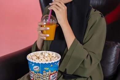 Enjoying refreshing drinks, locally produced Emirati products, a close-up portrait of an Arab Gulf Emirati woman wearing an abaya, holding popcorn and a juice cup in her hands, with a pink background.