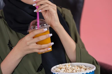 Local Emirati national products, enjoying refreshing drinks, a close-up portrait of an Arab Gulf Emirati woman wearing an abaya holding popcorn and a juice cup in her hands, with a pink background.