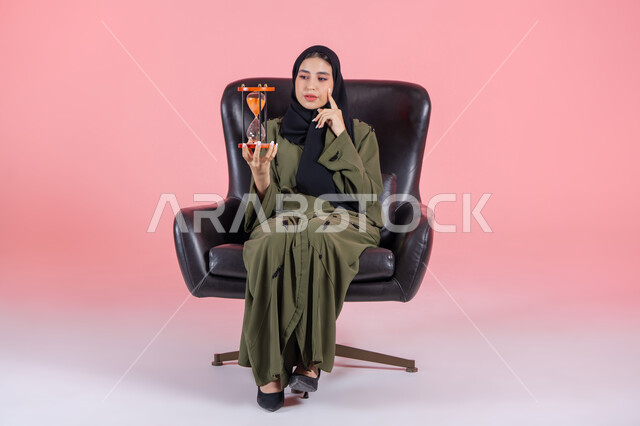 Reminder of a specific appointment, realizing the value of time and precious moments in our lives, the importance of punctuality and commitment to appointments, portrait of a veiled Emirati Gulf Arab woman wearing a green abaya sitting on a comfortable chair with her hand on her cheek and holding an hourglass, pink background