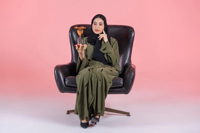 التذكير بموعد محدد, ادارك قيمة الوقت و اللحظات الثمينة في حياتنا, أهمية الدقة والالتزام في المواعيد, بورتريه لامرأة اماراتية خليجية عربية محجبة ترتدي عباءة خضراء تجلس على كرسي مريح و تضع يدها على خدها و تمسك ساعة رملية, خلفية وردية