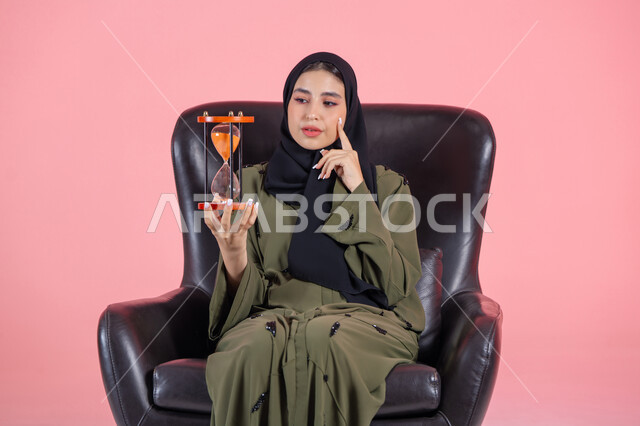 مفهوم تنظيم الوقت، الالتزام بالموعد المحدد في انجاز الاعمال، بورتريه لامرأة اماراتية خليجية عربية محجبة ترتدي عباءة خضراء تجلس على كرسي مريح و تضع يدها على خدها و تمسك ساعة رملية, خلفية وردية