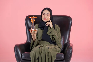 مفهوم تنظيم الوقت، الالتزام بالموعد المحدد في انجاز الاعمال، بورتريه لامرأة اماراتية خليجية عربية محجبة ترتدي عباءة خضراء تجلس على كرسي مريح و تضع يدها على خدها و تمسك ساعة رملية, خلفية وردية
