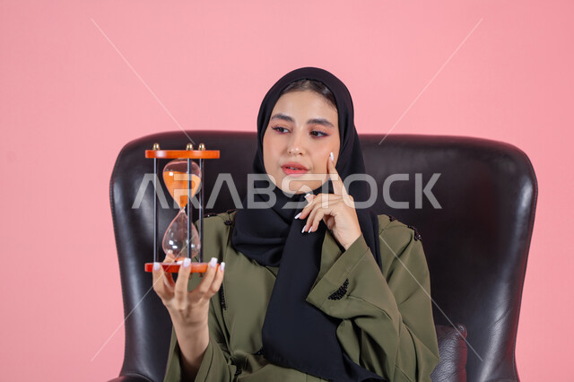 ادارك قيمة الوقت و اللحظات الثمينة في حياتنا, أهمية الدقة والالتزام في المواعيد, بورتريه لامرأة اماراتية خليجية عربية محجبة ترتدي عباءة خضراء تجلس على كرسي مريح و تضع يدها على خدها و تمسك ساعة رملية, خلفية وردية