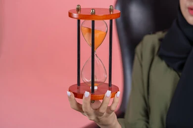 الدقة في المواعيد, صوره مقربه ليد امرأة اماراتية خليجية عربية محجبة ترتدي عباءة خضراء تجلس على كرسي مريح و تمسك ساعه رمليه, إدارة الأعمال و تنظيم الجدول الزمني, القيام بمراقبة توقيت الرمل, إدراك قيمة اللحظات الثمينة, خلفية وردية